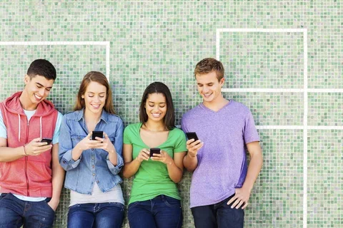 Group of four individuals standing against a green tile background holding their phones and smiling at them.